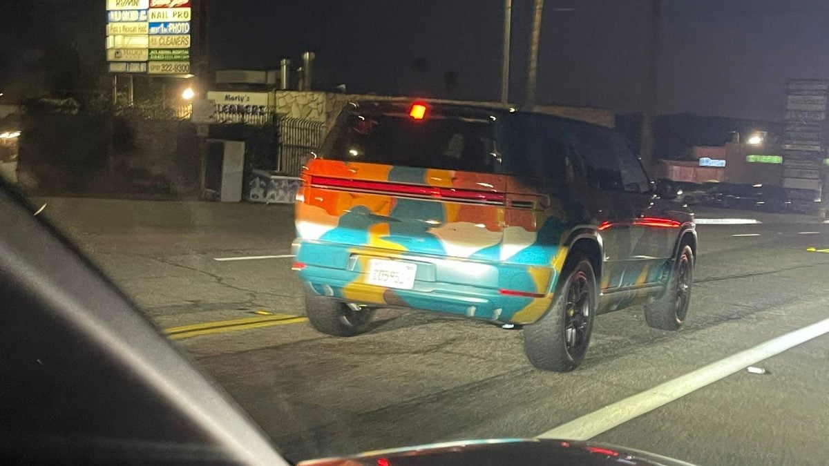 2026 Rivian R2 electric SUV rear three-quarter view in multi-color camouflage wrap driving on urban road at night.